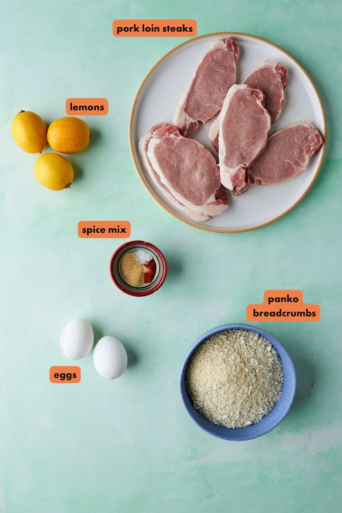 Flat lay of raw pork loin steaks on a plate, three lemons, a small bowl of spice mix, two eggs, and a bowl of panko breadcrumbs with roasted corn, all labeled on a green background.