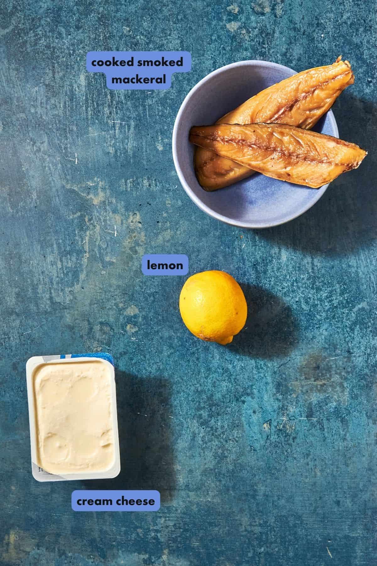 A bowl with two pieces of cooked smoked mackerel, roasted corn, a whole lemon, and a container of cream cheese are arranged on a blue surface with labeled text indicating each item.