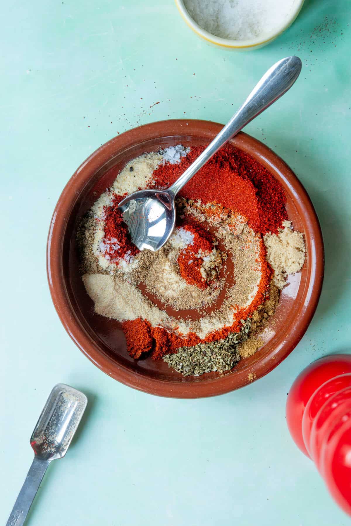 A brown bowl with various colorful ground spices, including red, white, and green powders arranged in swirls, sits alongside a metal measuring spoon and small bowl on a light blue surface. Roasted corn seasoning adds a unique touch to the mix.