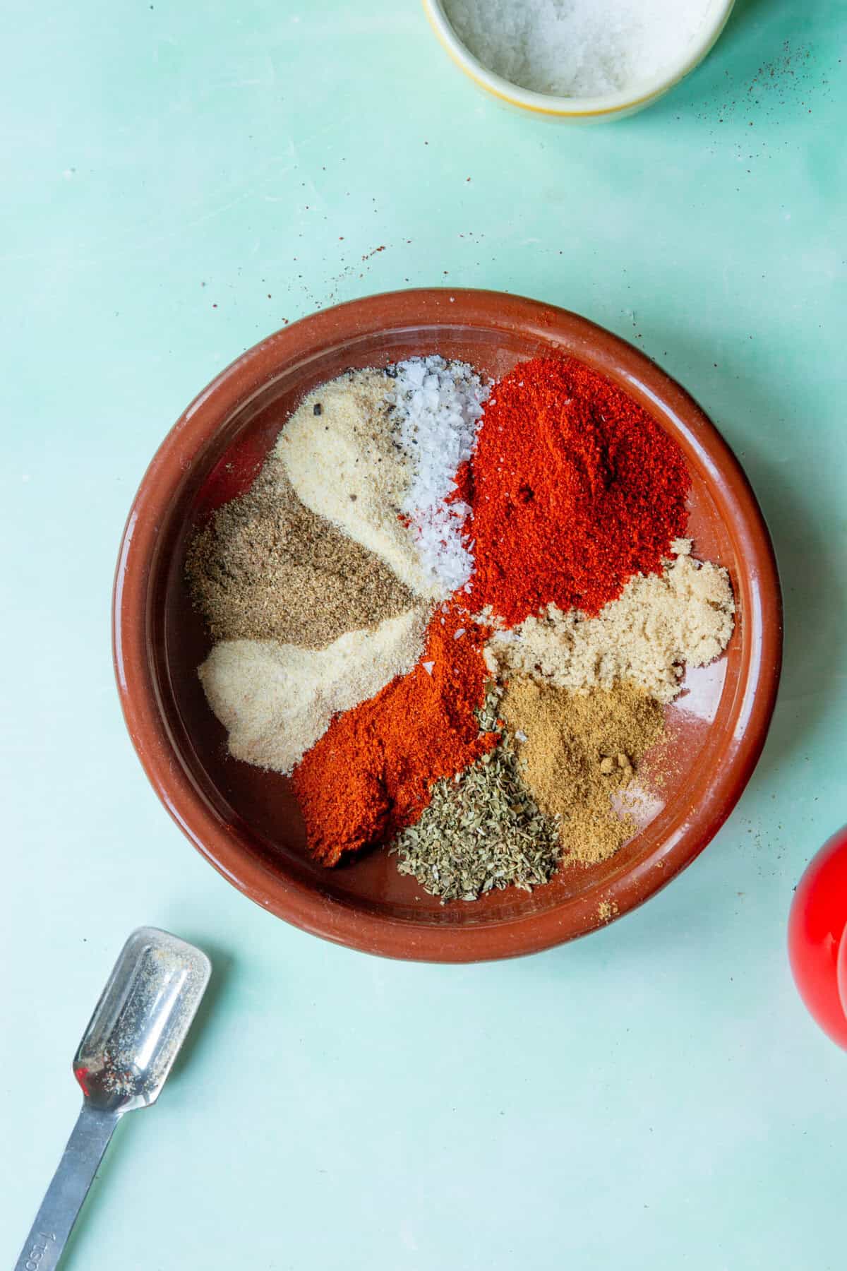 A terracotta bowl filled with various colorful ground spices and herbs, including roasted corn powder alongside red, brown, white, and green piles, sits on a pale green surface with a metal measuring spoon nearby.