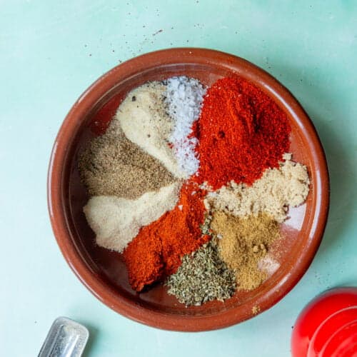 A brown ceramic bowl contains mounds of colorful spices, including red paprika, salt, dried herbs, and ground seasonings—perfect for flavoring roasted corn—arranged separately on a light blue surface. A metal scoop and a red item are nearby.
