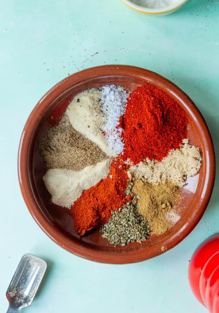 A brown ceramic bowl contains mounds of colorful spices, including red paprika, salt, dried herbs, and ground seasonings—perfect for flavoring roasted corn—arranged separately on a light blue surface. A metal scoop and a red item are nearby.