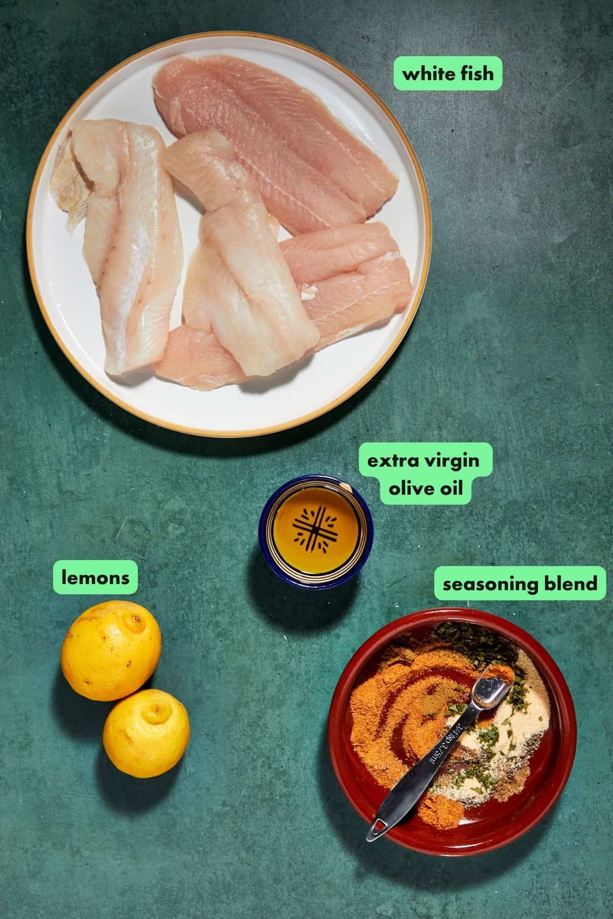 A bowl of raw white fish fillets, two whole lemons, a glass of extra virgin olive oil, roasted corn, and a bowl of mixed seasonings on a green surface, each ingredient labeled with text.