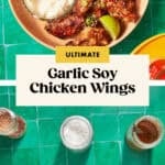 A bowl of soy chicken wings with dipping sauce and lime wedges sits above ingredients like raw chicken wings, flour, seasonings, and sauce jars on a green tile surface. Text: “Ultimate Garlic Soy Chicken Wings.”.