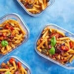 Four glass containers filled with vibrant Mediterranean Pasta Salad, garnished with basil leaves, are arranged on a blue surface. Text highlights this quick meal prep recipe—Mediterranean Pasta Salad serves 5 and is ready in just 35 minutes.