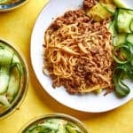 A plate of Peanut Sauce Noodles with ground meat and sliced cucumber sits on a yellow background, surrounded by three bowls of cucumber noodles. Text reads 15 Minute Peanut Noodles, High Protein Noodle Bowls, and www.beatthebudget.com.