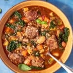 Overhead view of a bowl of Beef Minestrone Soup with vegetables and herbs on a blue background, spoon in the bowl. Text reads Best Beef Minestrone Soup and Easy Fuss-Free Family Dinner.