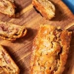 A sliced bacon roly poly pastry, reminiscent of classic bacon pudding, sits on a round wooden board with a golden crust and visible bacon pieces inside. Text reads Bacon Roly Poly and www.beatthebudget.com on a blue background.