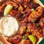 A plate of soy chicken wings glazed in garlic and garnished with lime wedges and green onions, served with creamy dipping sauce on a green tiled surface. Text reads Garlic Soy Chicken Wings and www.beatthebudget.com.