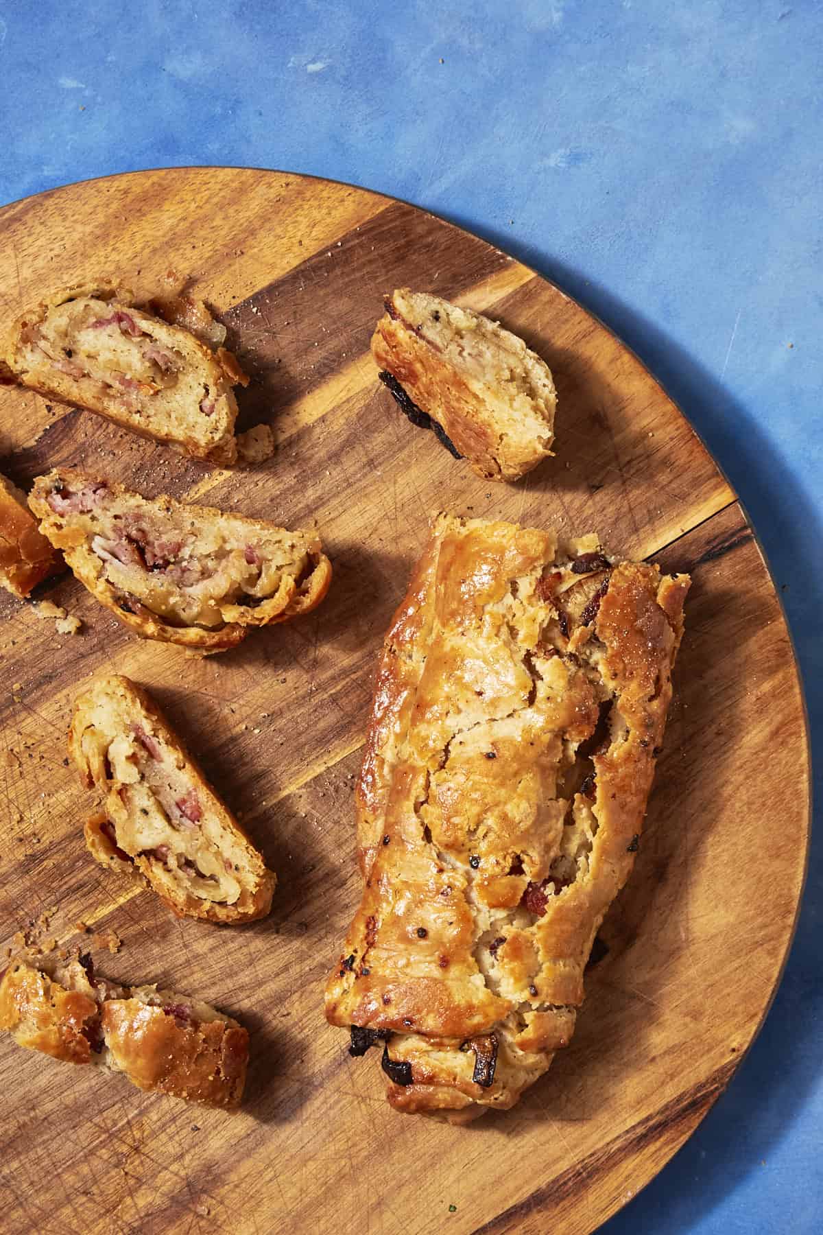 A sliced, golden-brown savory pastry with a flaky crust and visible bacon pudding filling, arranged on a round wooden board against a bright blue background.