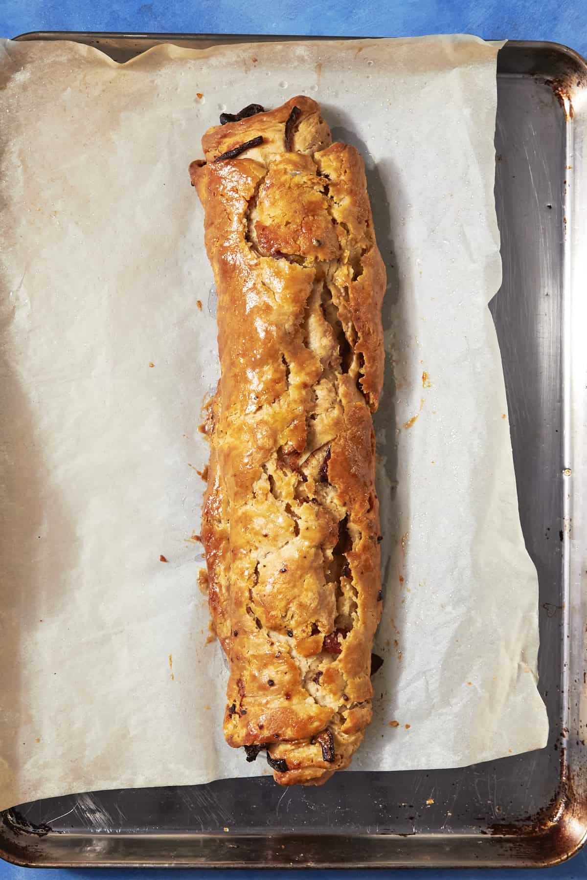 A golden-brown, baked strudel sits on a sheet of parchment paper on a metal baking tray, its cracked and flaky crust hinting at a savory bacon pudding filling inside.