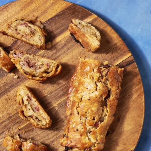 A golden-brown pastry roll, partially sliced, sits on a round wooden board against a blue background. Several pieces reveal a filling reminiscent of bacon pudding with nuts and possibly fruit inside the flaky crust.