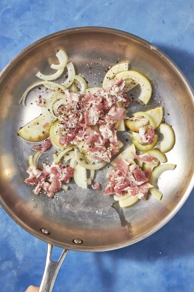 Sliced onions and raw bacon for bacon pudding cooking in a stainless steel skillet, seasoned with black pepper, on a blue countertop.