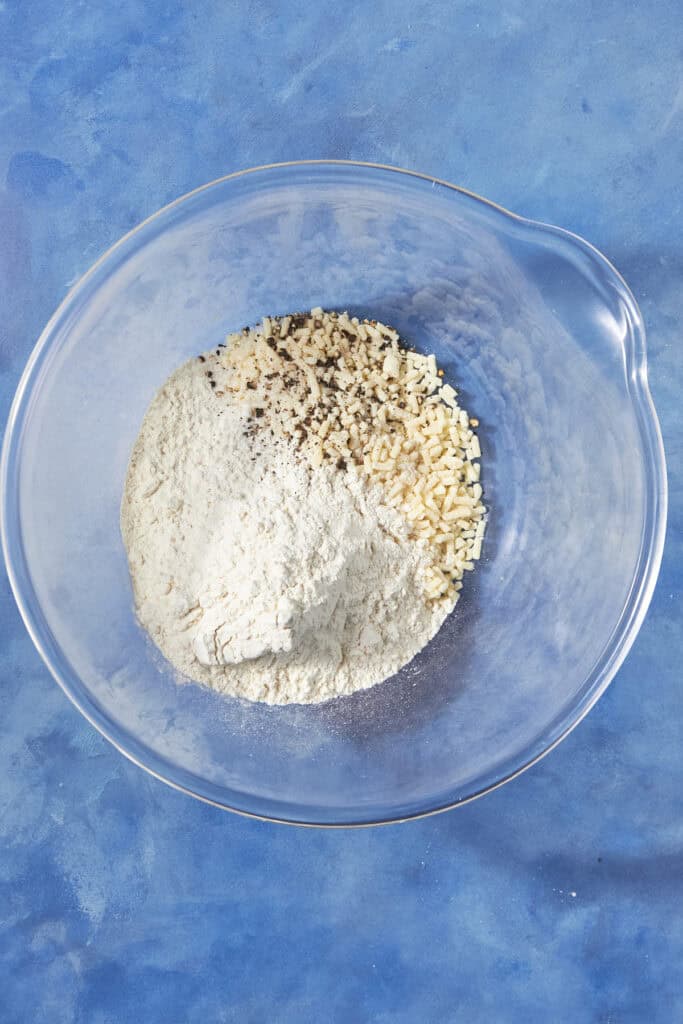 A clear glass mixing bowl on a blue surface contains flour and minced dried onions with some black pepper sprinkled on top, ready to become a savory bacon pudding.
