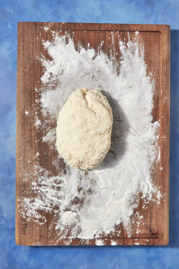 A ball of dough sits on a wooden cutting board dusted with flour, ready to be transformed into a savory bacon pudding, against a blue background.