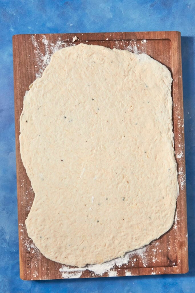 A large, unevenly rolled piece of pizza dough, waiting to be topped with bacon pudding, rests on a floured wooden board against a blue background.
