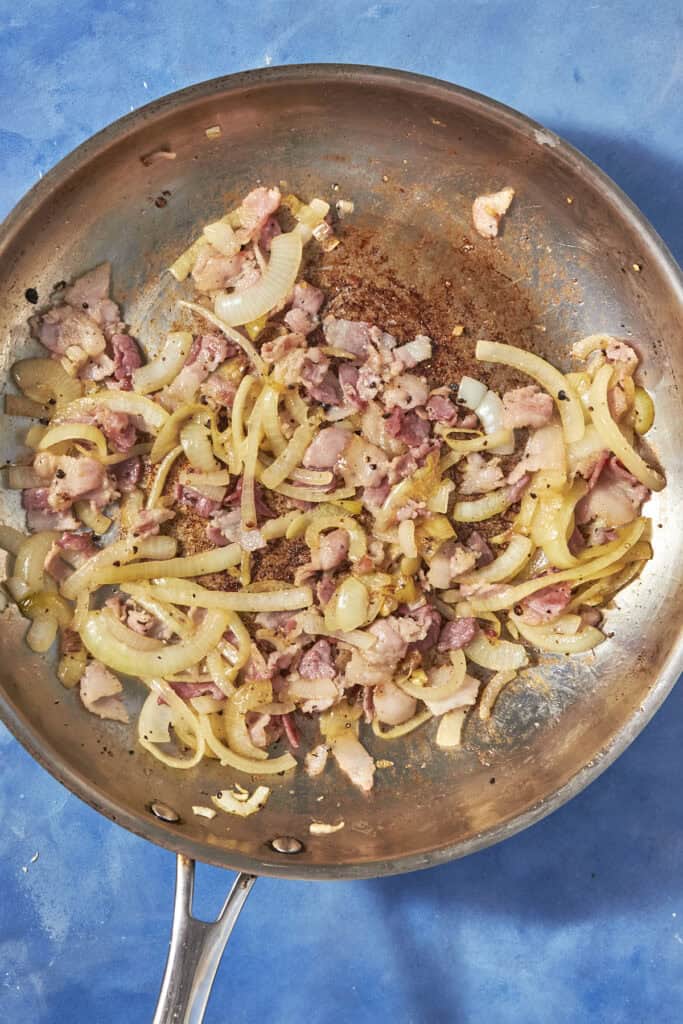 A stainless steel skillet on a blue surface contains sautéed onions and pieces of bacon, with browned bits and some oil visible—perfect savory ingredients for a classic bacon pudding.