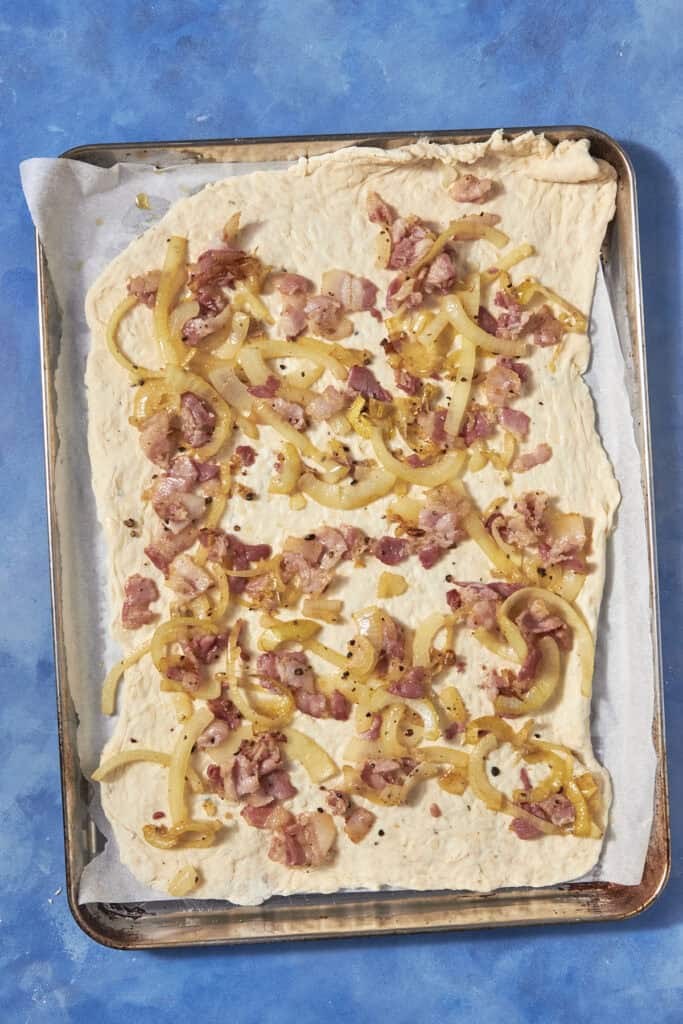 Rectangular sheet of raw dough on a baking tray lined with parchment paper, topped with cooked bacon pieces and sautéed onions—perfect for a savory twist on classic bacon pudding—set against a blue background.