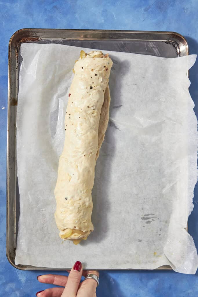 A hand holds a baking tray lined with parchment paper, topped with an uncooked bacon pudding dough log, ready for baking. The background is a bright blue surface.