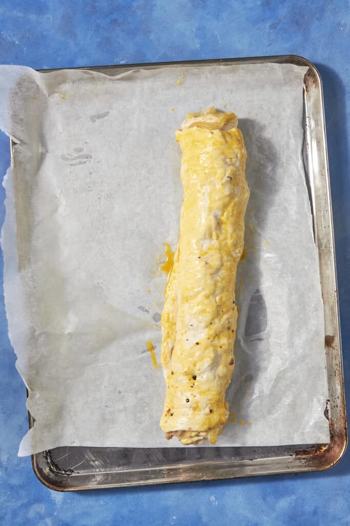 A rolled pastry, reminiscent of classic bacon pudding, sits on a parchment-lined baking tray covered in egg wash, ready to be baked against a bright blue background.