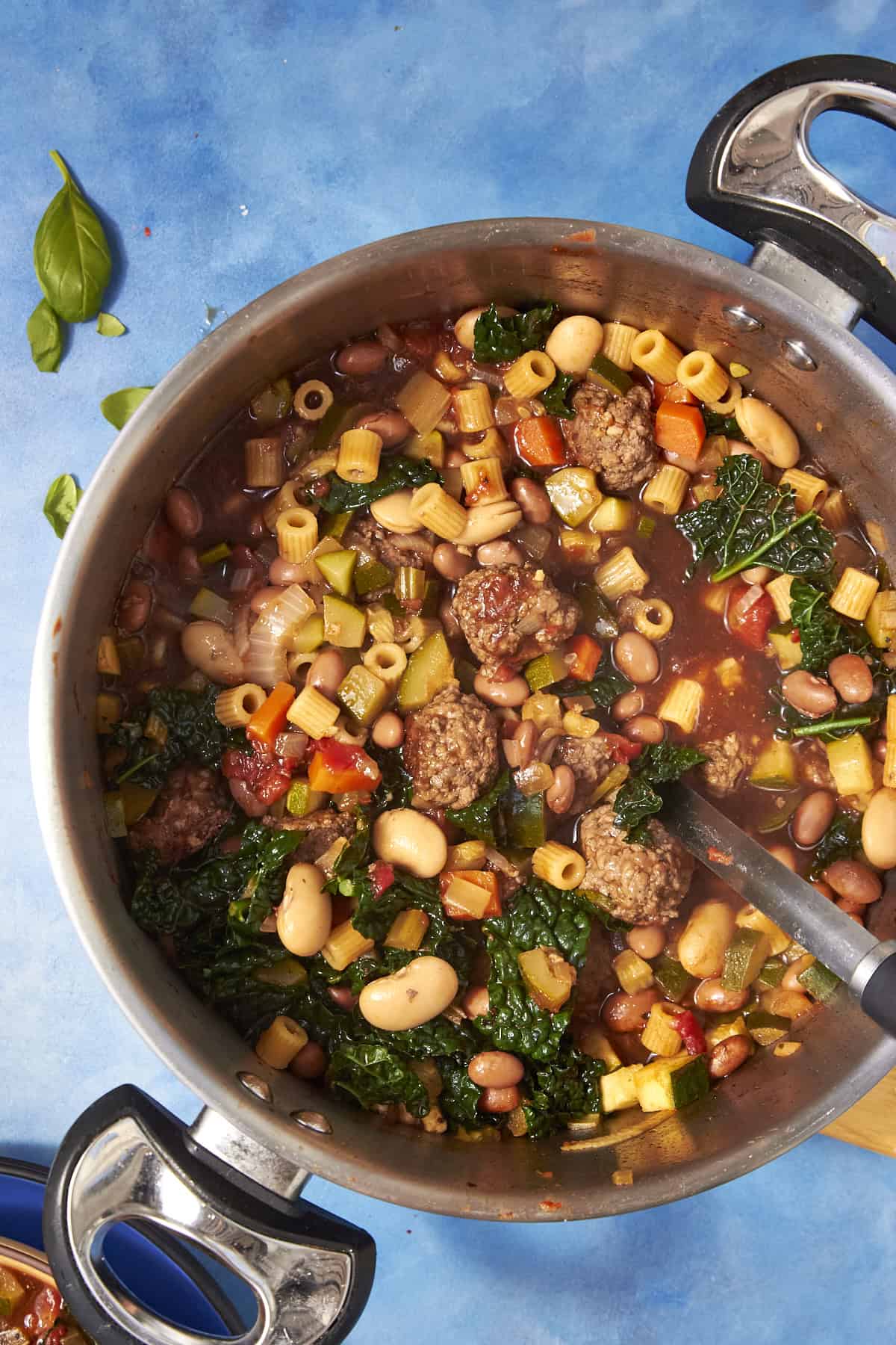 A large pot of hearty Beef Minestrone Soup with meatballs, pasta, beans, kale, and vegetables in a tomato-based broth sits on a blue surface with a few basil leaves nearby.