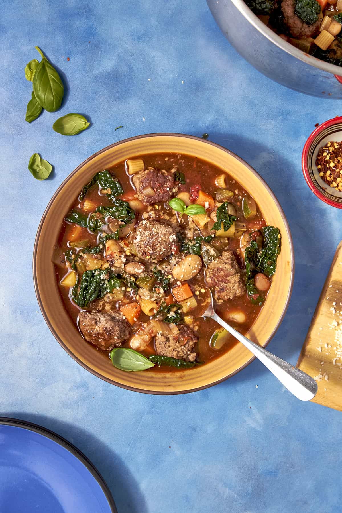A bowl of hearty Beef Minestrone Soup with meatballs, beans, pasta, kale, and vegetables sits on a blue surface. A spoon rests in the bowl, with basil leaves and chili flakes nearby.