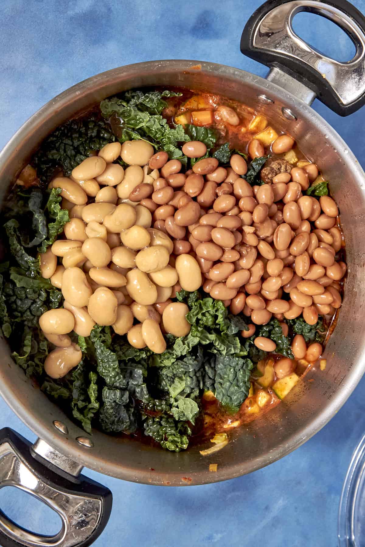A pot filled with dark green kale, lima beans, and pinto beans atop a tomato-based Beef Minestrone Soup, seen from above on a blue surface.