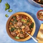 A bowl of hearty Beef Minestrone Soup with meatballs, beans, leafy greens, and chopped vegetables on a blue surface. Fresh basil leaves and a pot of soup are nearby, along with a small bowl of red pepper flakes.