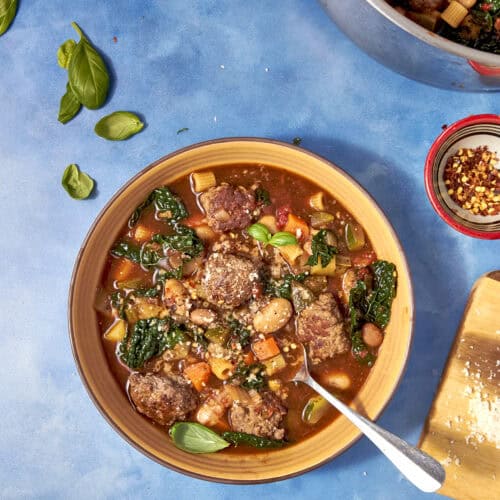 A bowl of hearty Beef Minestrone Soup with meatballs, beans, leafy greens, and chopped vegetables on a blue surface. Fresh basil leaves and a pot of soup are nearby, along with a small bowl of red pepper flakes.
