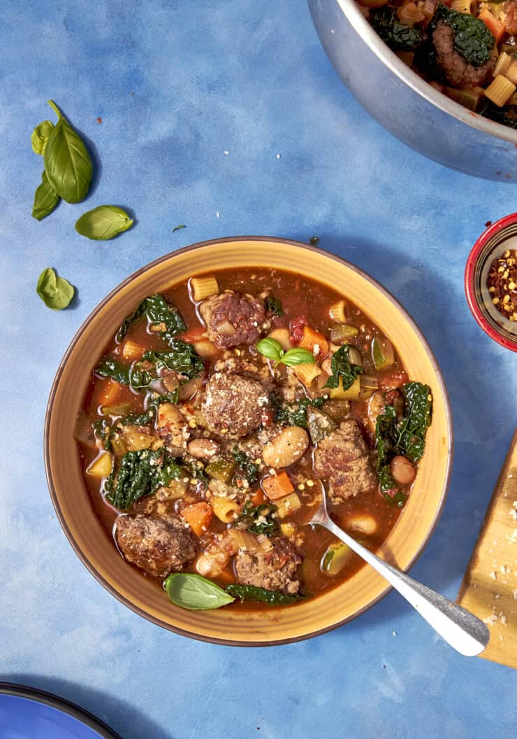 A bowl of hearty Beef Minestrone Soup with meatballs, beans, leafy greens, and chopped vegetables on a blue surface. Fresh basil leaves and a pot of soup are nearby, along with a small bowl of red pepper flakes.