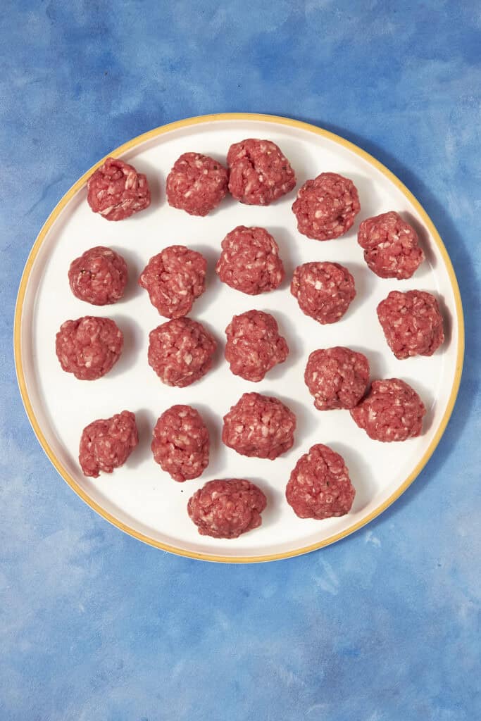 A white plate with a yellow rim holds 21 raw meatballs arranged in rows, ready to be added to Beef Minestrone Soup, placed on a blue textured surface.