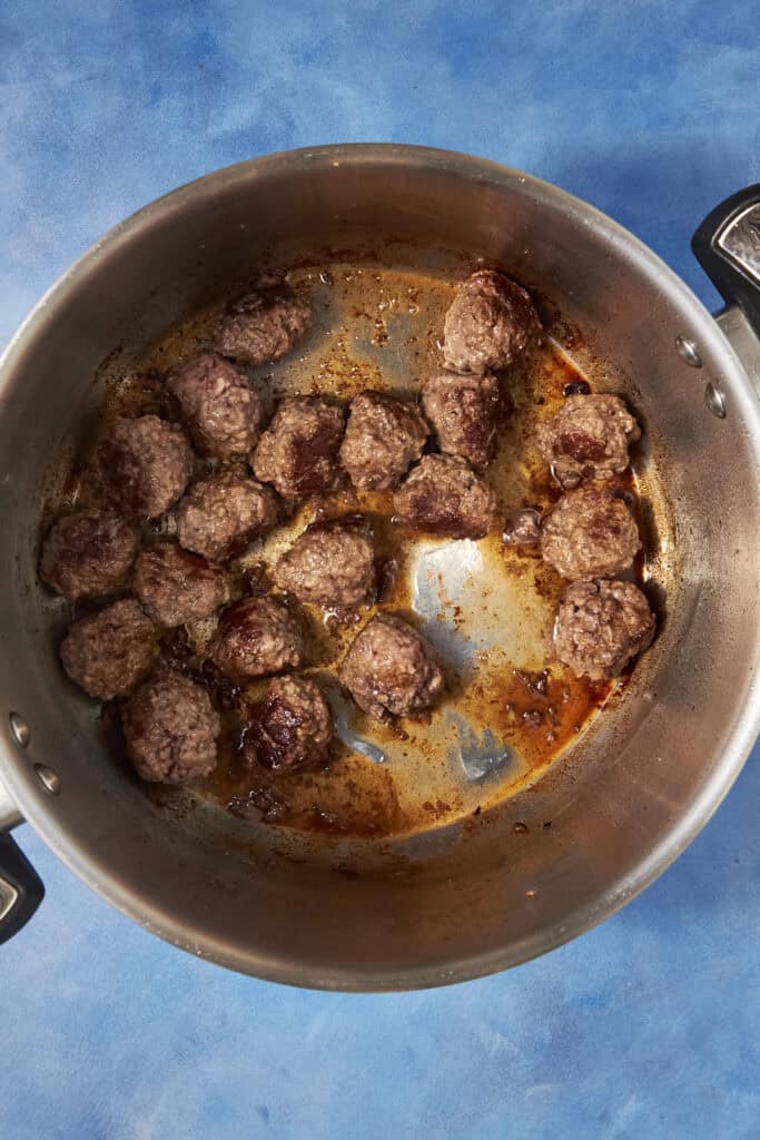 A large metal pot containing browned meatballs and some oil on the bottom, ready to add to a hearty Beef Minestrone Soup, set against a blue background.
