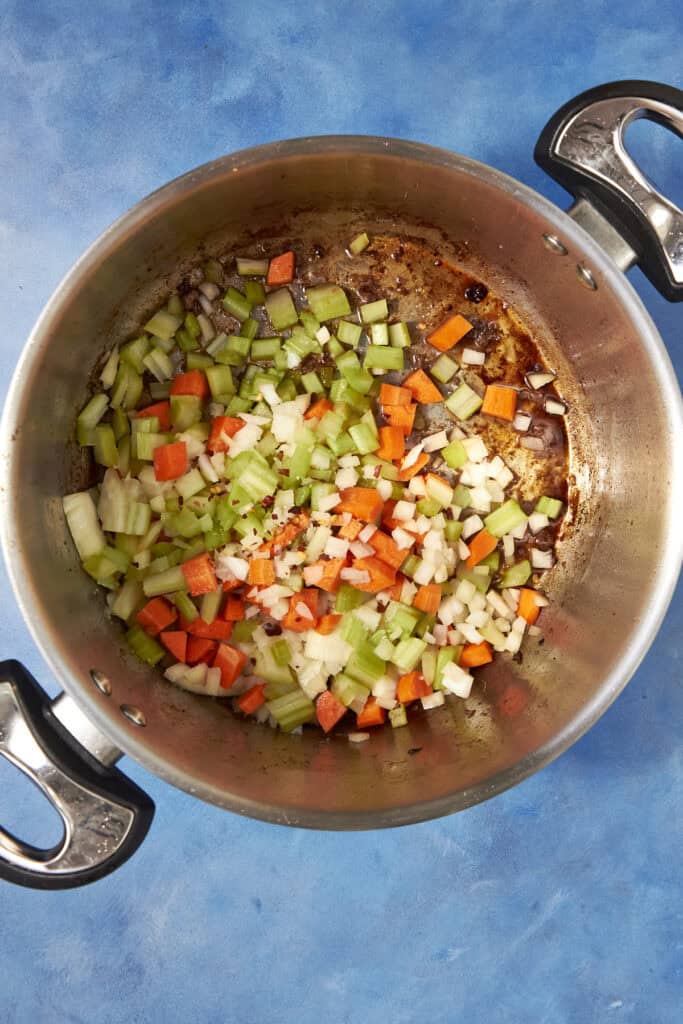 A large pot on a blue surface contains chopped onions, celery, and carrots beginning to sauté for Beef Minestrone Soup, with bits of browned fond visible on the bottom of the pot.