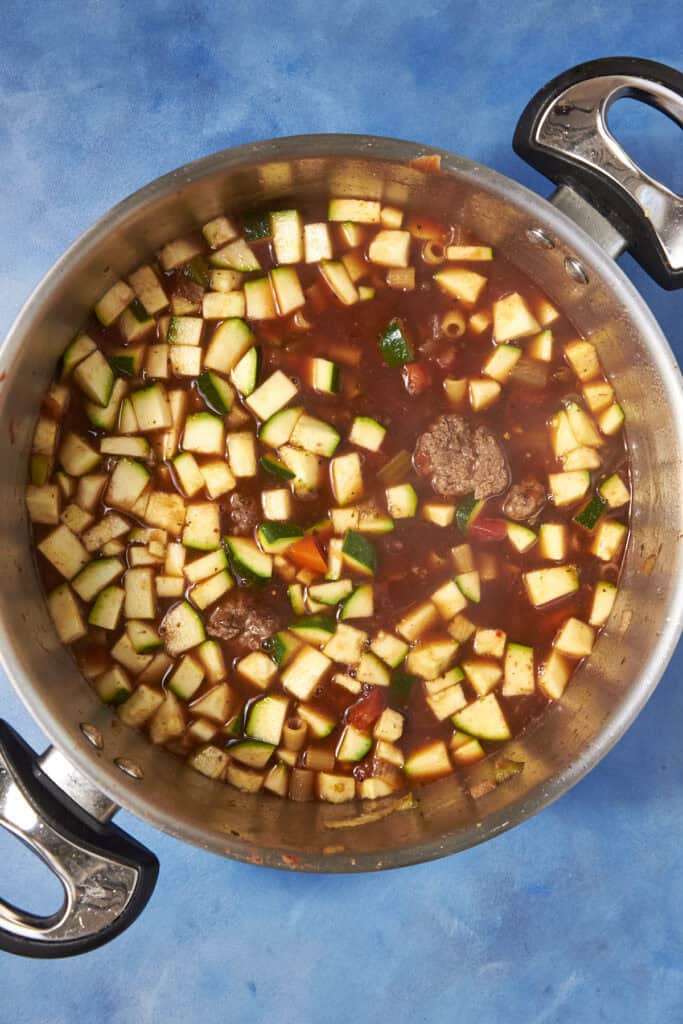 A large pot filled with chunky Beef Minestrone Soup, featuring diced zucchini, carrots, and meatballs in a savory broth, sits on a blue surface.