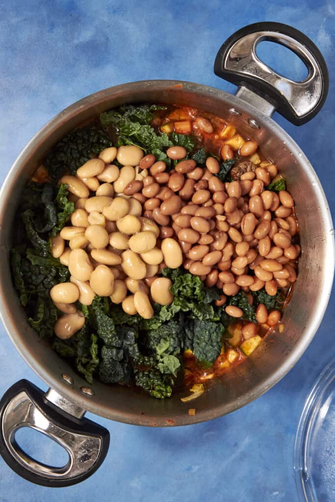 A pot on a blue surface filled with kale, white beans, and pinto beans, arranged in sections and partially mixed with a hearty Beef Minestrone Soup tomato-based stew.