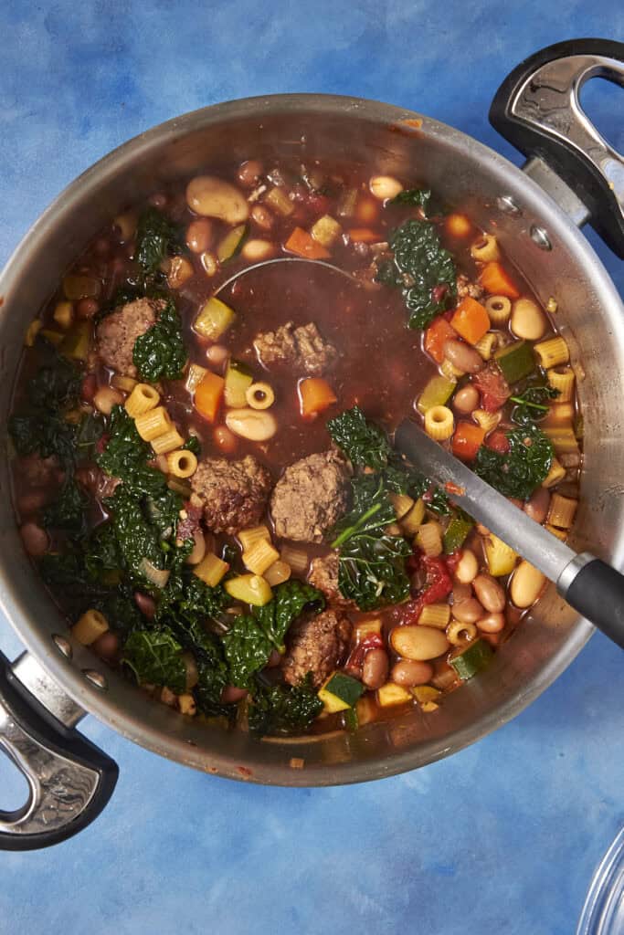 A pot of hearty Beef Minestrone Soup with meatballs, pasta, white beans, greens, carrots, zucchini, and tomatoes in a rich broth, with a ladle resting inside the pot on a blue surface.
