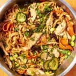 A bowl of noodle salad with shredded chicken, sliced cucumber, carrot ribbons, edamame, roasted corn, herbs, and a dark sauce, all mixed together on a bright orange background.