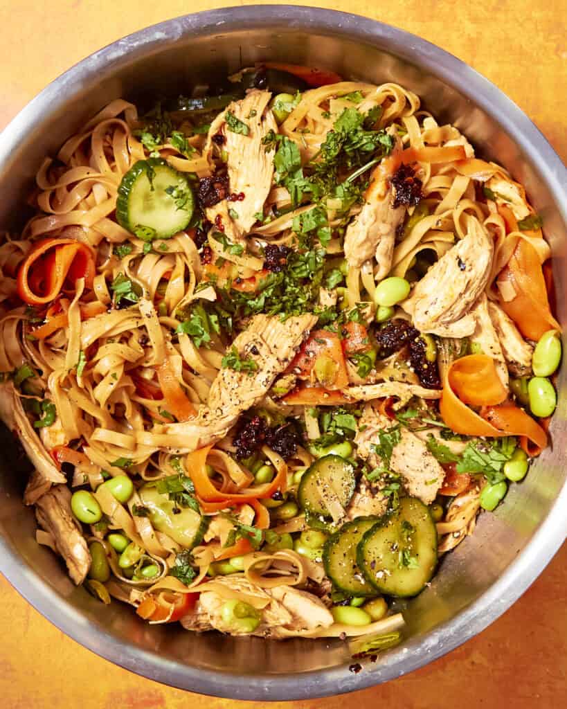 A bowl of noodle salad with shredded chicken, sliced cucumber, carrot ribbons, edamame, roasted corn, herbs, and a dark sauce, all mixed together on a bright orange background.