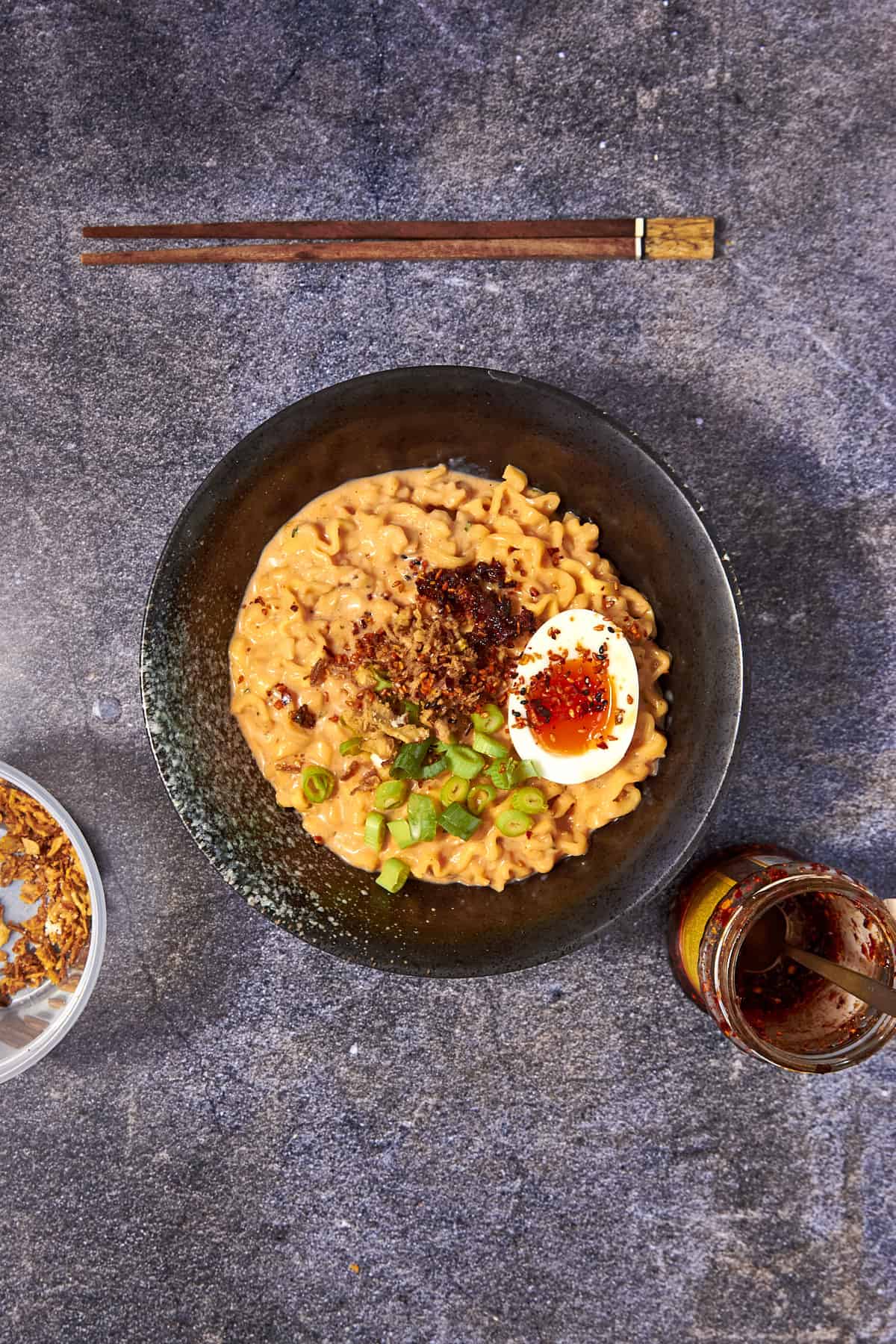 A bowl of creamy buldak noodles topped with a halved soft-boiled egg, chopped green onions, and chili oil sits on a dark surface with chopsticks above and a jar of chili oil to the right.