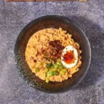 A bowl of creamy buldak noodles topped with a halved soft-boiled egg, chopped green onions, chili flakes, and fried onions, with wooden chopsticks placed above the bowl on a stone surface.