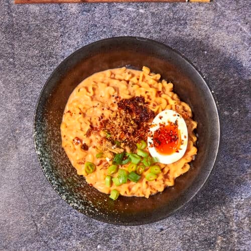 A bowl of creamy buldak noodles topped with a halved soft-boiled egg, chopped green onions, chili flakes, and fried onions, with wooden chopsticks placed above the bowl on a stone surface.
