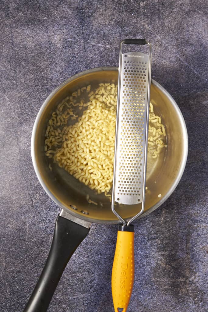 A saucepan with grated cheese, perfect for creamy buldak noodles, sits on a textured countertop. A metal cheese grater with an orange handle rests across the pan.