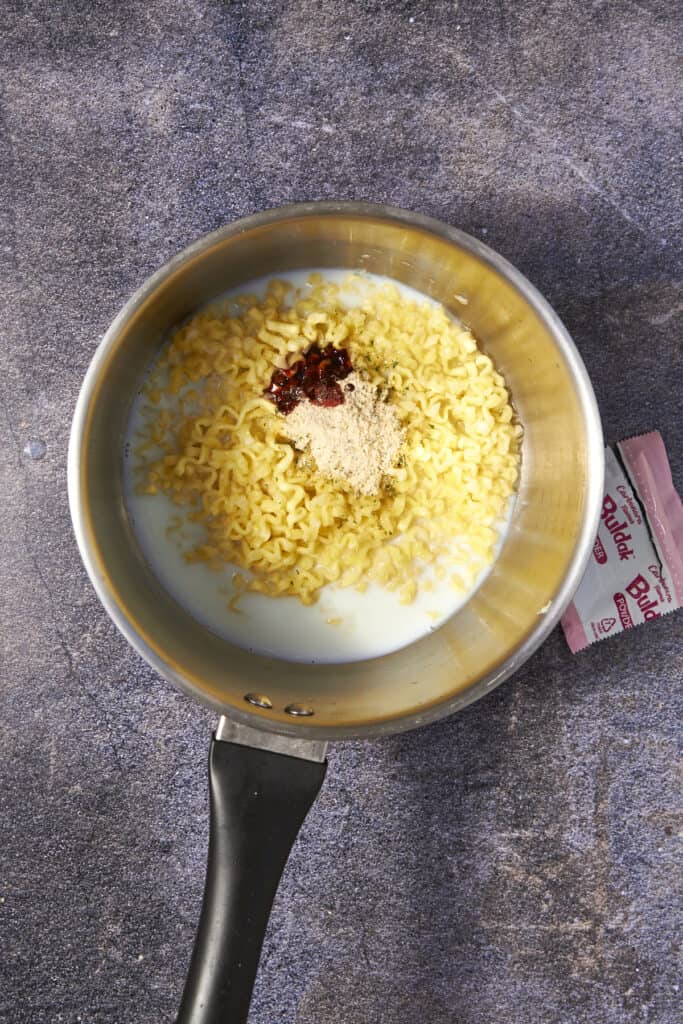 A saucepan with creamy buldak noodles, dried ramen, milk, soup base powder, and chili paste sits on a dark countertop. An empty ramen seasoning packet is partially visible next to the pan.