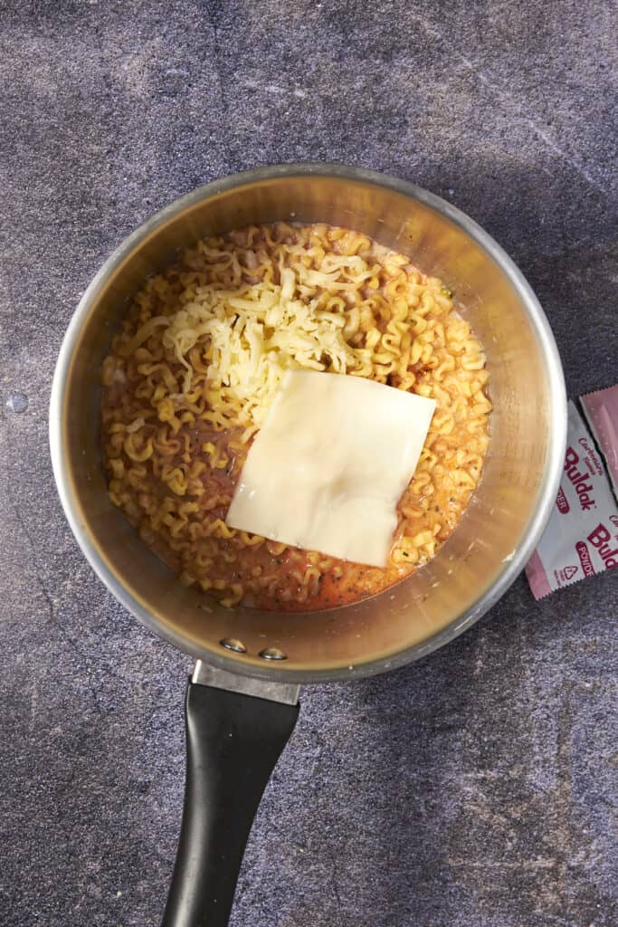 A pot of cooked creamy buldak noodles topped with a slice of processed cheese and shredded cheese sits on a textured gray surface. An opened seasoning packet is partially visible to the side.