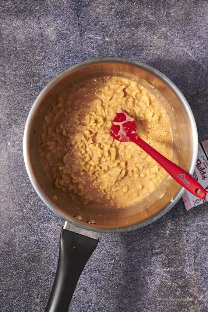 A saucepan filled with creamy buldak noodles in a rich, orange cheese sauce, stirred with a red spatula. The pot rests on a dark textured surface, with a partially visible seasoning packet beside it.