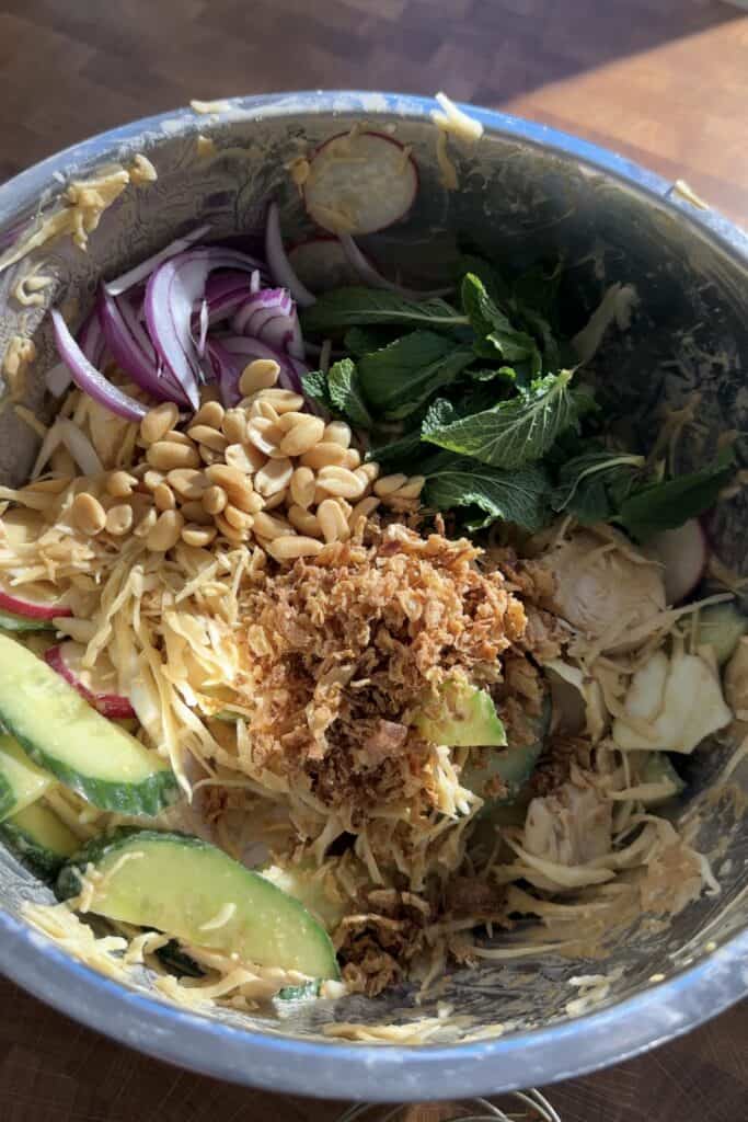 A metal bowl holds a vibrant Peanut Chicken Cabbage Salad with shredded chicken, cucumber slices, red onion strips, radish, mint leaves, peanuts, and crispy fried shallots arranged beautifully on a wooden surface.