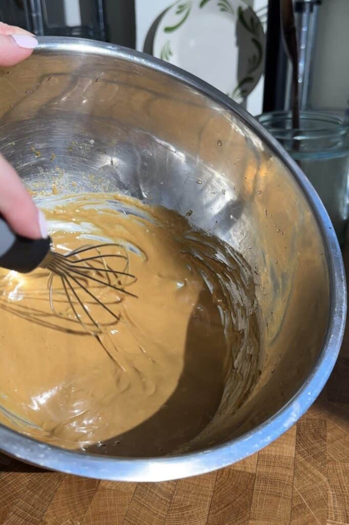 A hand holding a whisk mixes a thick, light brown batter for Peanut Chicken Cabbage Salad in a shiny metal bowl on a wooden surface, with sunlight casting shadows inside the bowl.