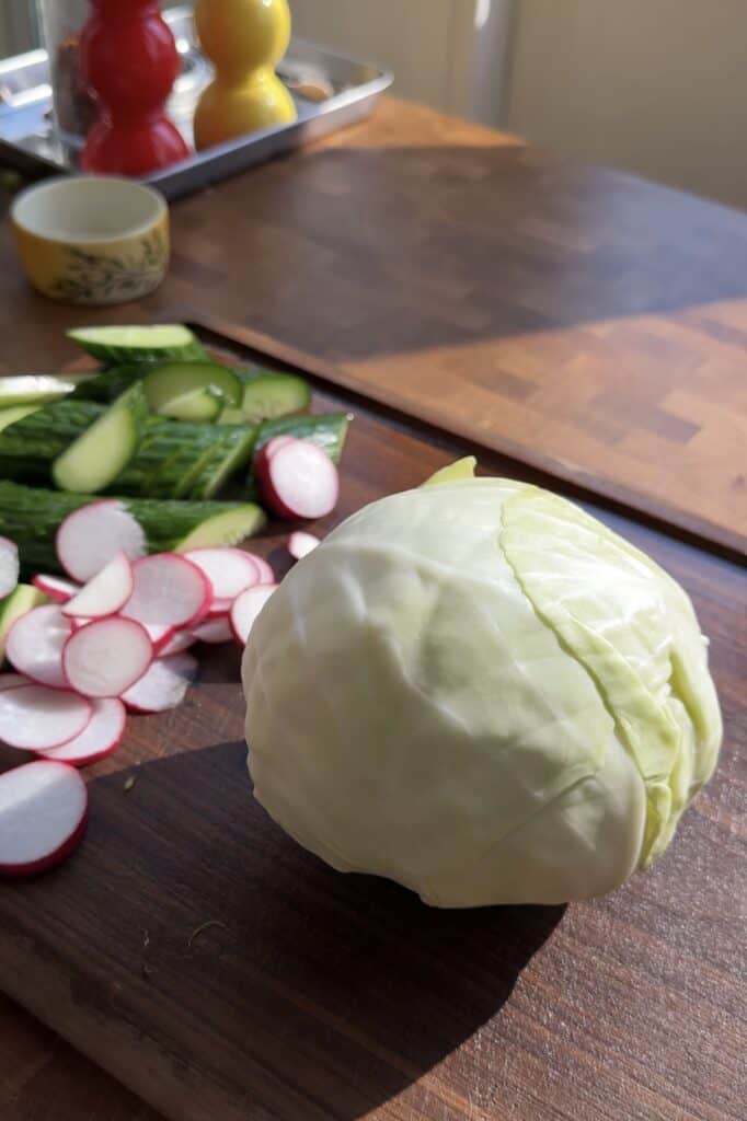 A head of cabbage sits on a wooden cutting board next to slices of cucumber and radish—fresh ingredients perfect for making Peanut Chicken Cabbage Salad. Sunlight shines on the vegetables, with other kitchen items blurred in the background.