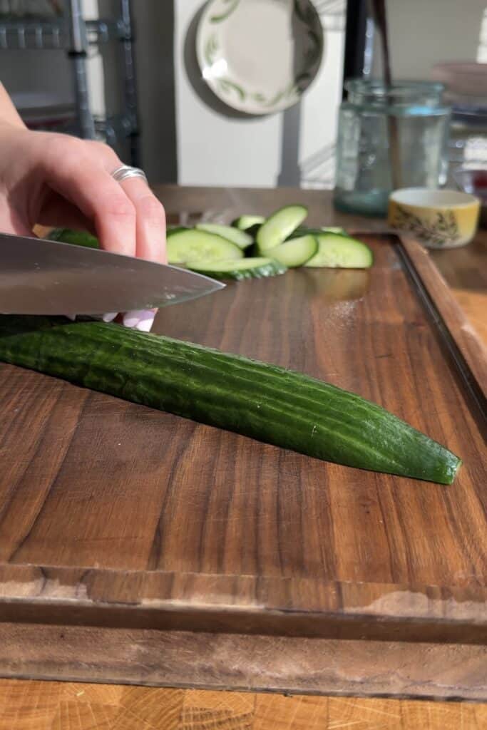 A hand is slicing a cucumber with a knife on a wooden cutting board, prepping ingredients for Peanut Chicken Cabbage Salad. Sliced cucumber pieces sit in the background next to a cup and dishes as sunlight shines on the scene.