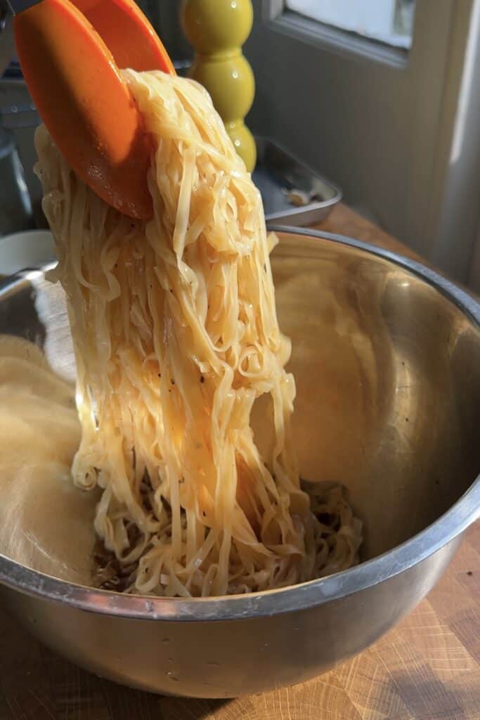 A pair of orange tongs lifts cooked noodles coated in sauce and roasted corn from a stainless steel mixing bowl on a wooden countertop, with sunlight streaming through a nearby window.